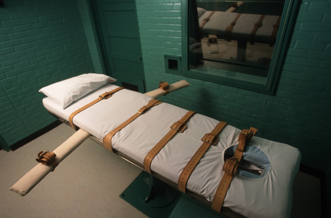 An execution chamber features a lethal injection gurney fitted with leather restraint straps and a white pillow, set against green brick walls.