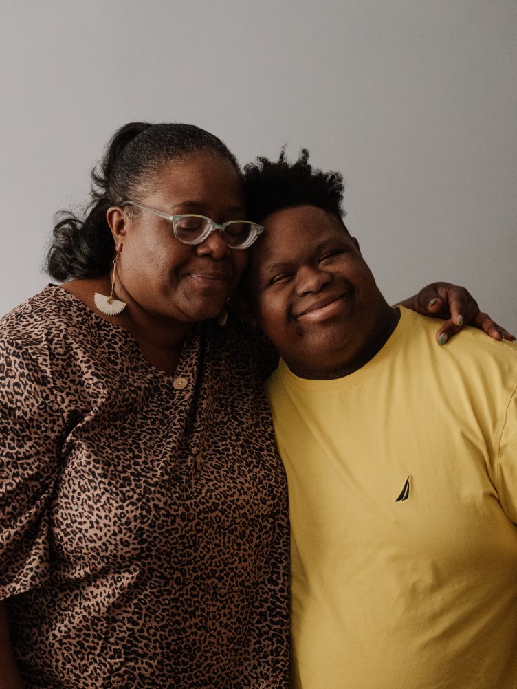 A mother, wearing glasses and a leopard-print shirt, and son, wearing a bright yellow shirt, smile and pose with their heads leaning together. The mother’s eyes are closed and the son is smiling.