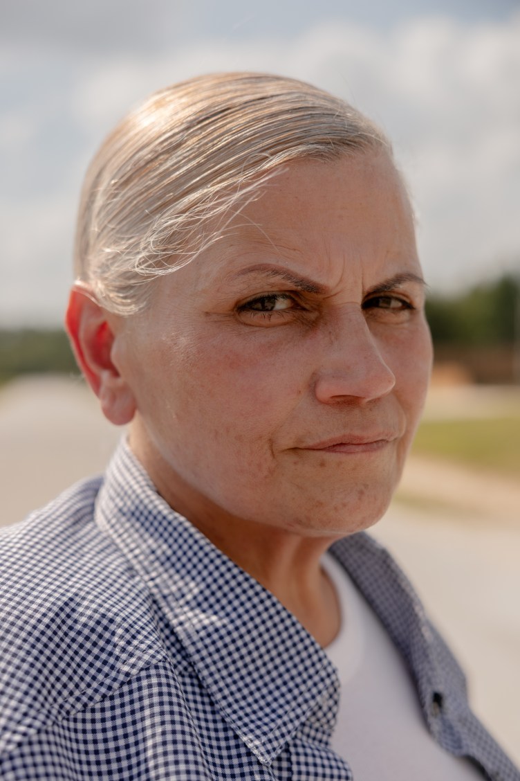 A woman with white hair, wearing a blue-checkered collared shirt over a white T-shirt, stares with a thin smile.