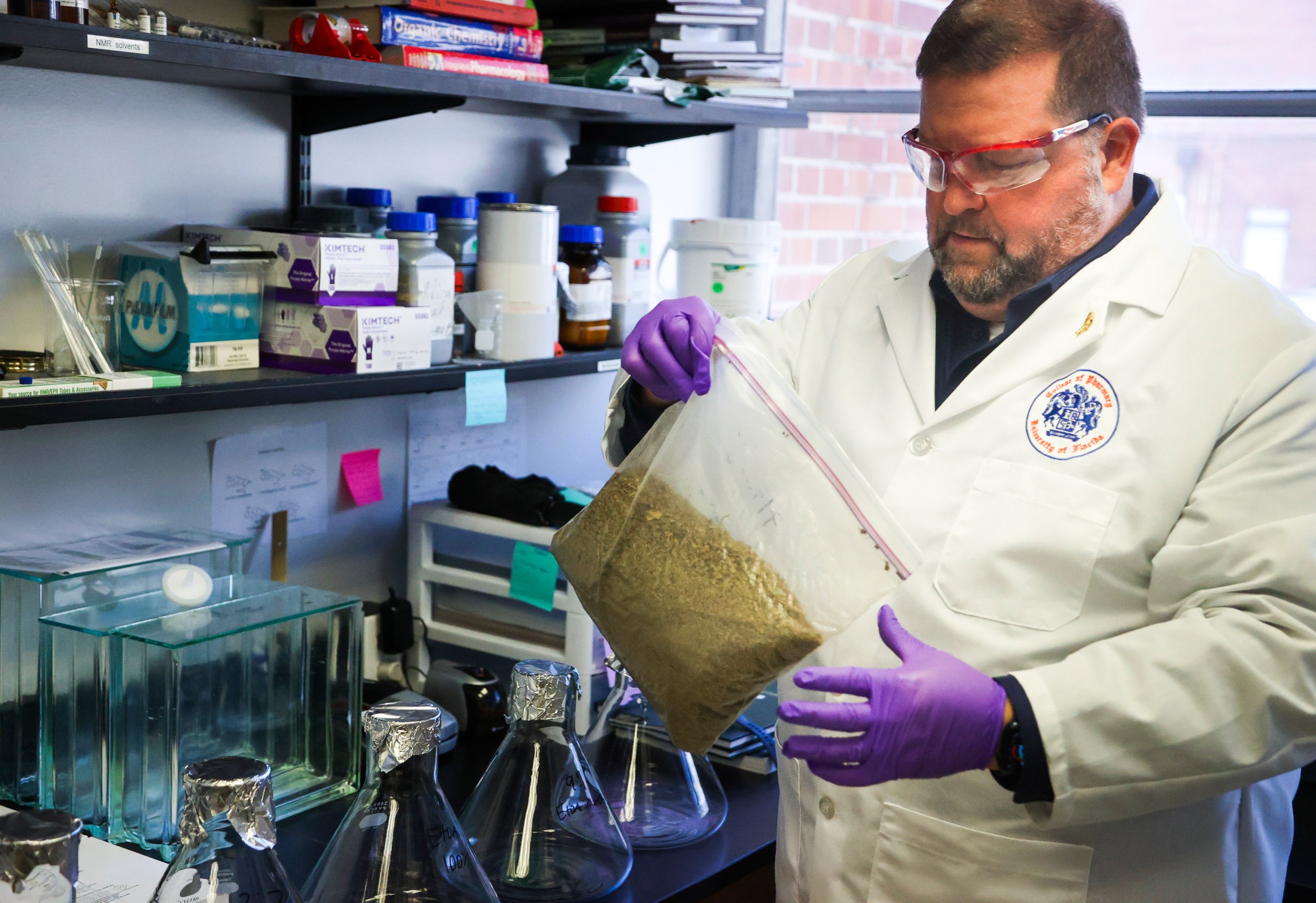 A bearded man in a lab coat handles a large Ziploc bag full of dried and ground-up plant material.