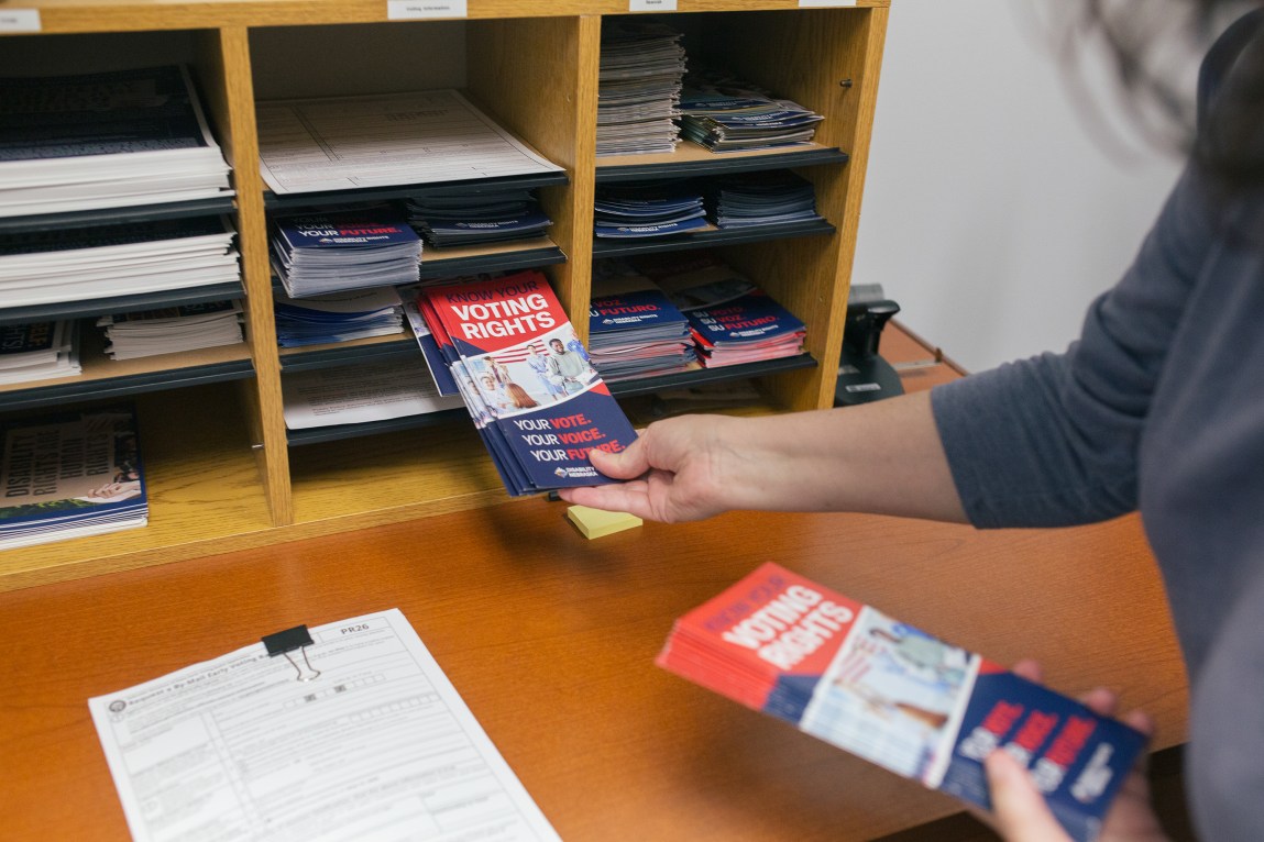 A woman’s arm and hand are seen placing a stack of red, white and blue pamphlets into a tabletop shelving unit.