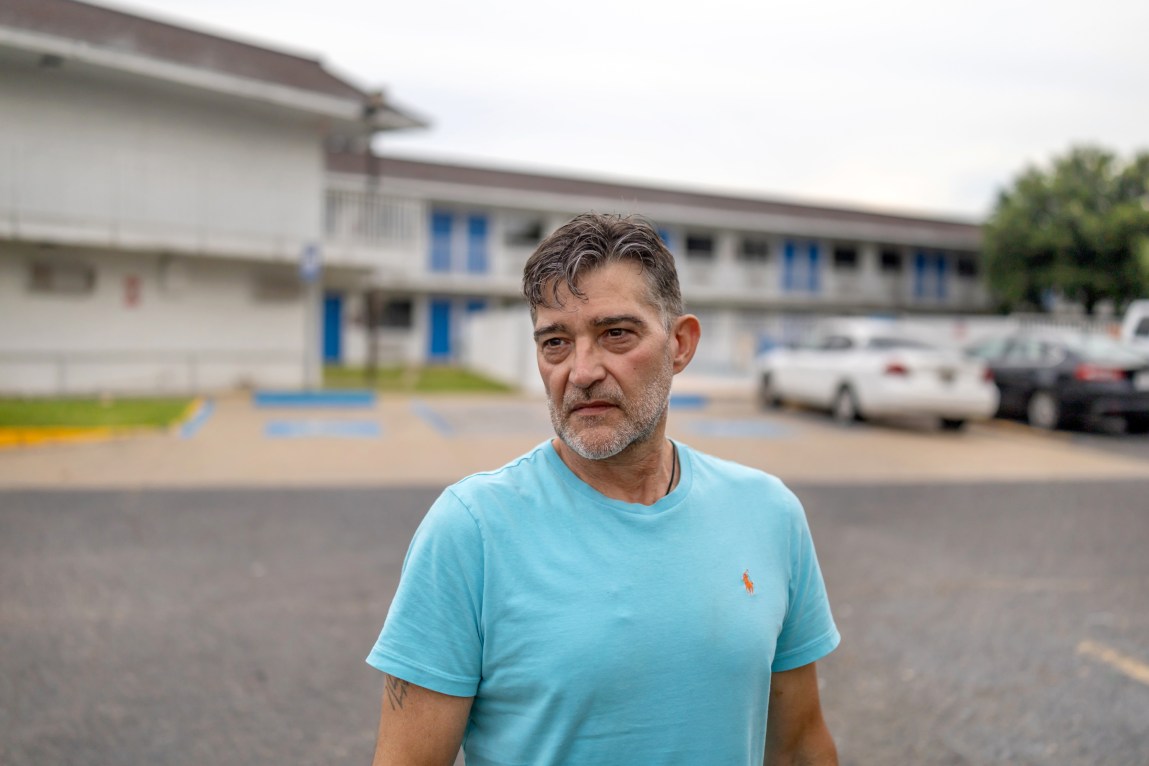 A man with graying hair and stubble looks off camera with a serious expression. Behind him is a parking lot and a motel building.