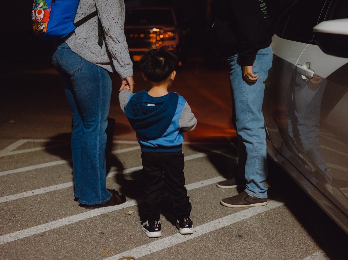 A young boy in a blue and gray hoodie stands in a parking lot at night, holding the hand of an adult in jeans and a gray sweater on the left. Another person in jeans stands on the right next to a white car.