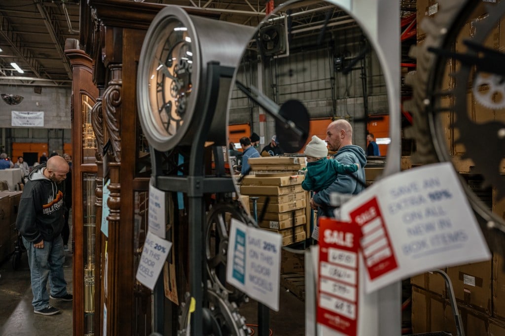 One man leans down to closely examine a grandfather clock, while another man is reflected in the mirrored face of another nearby clock.