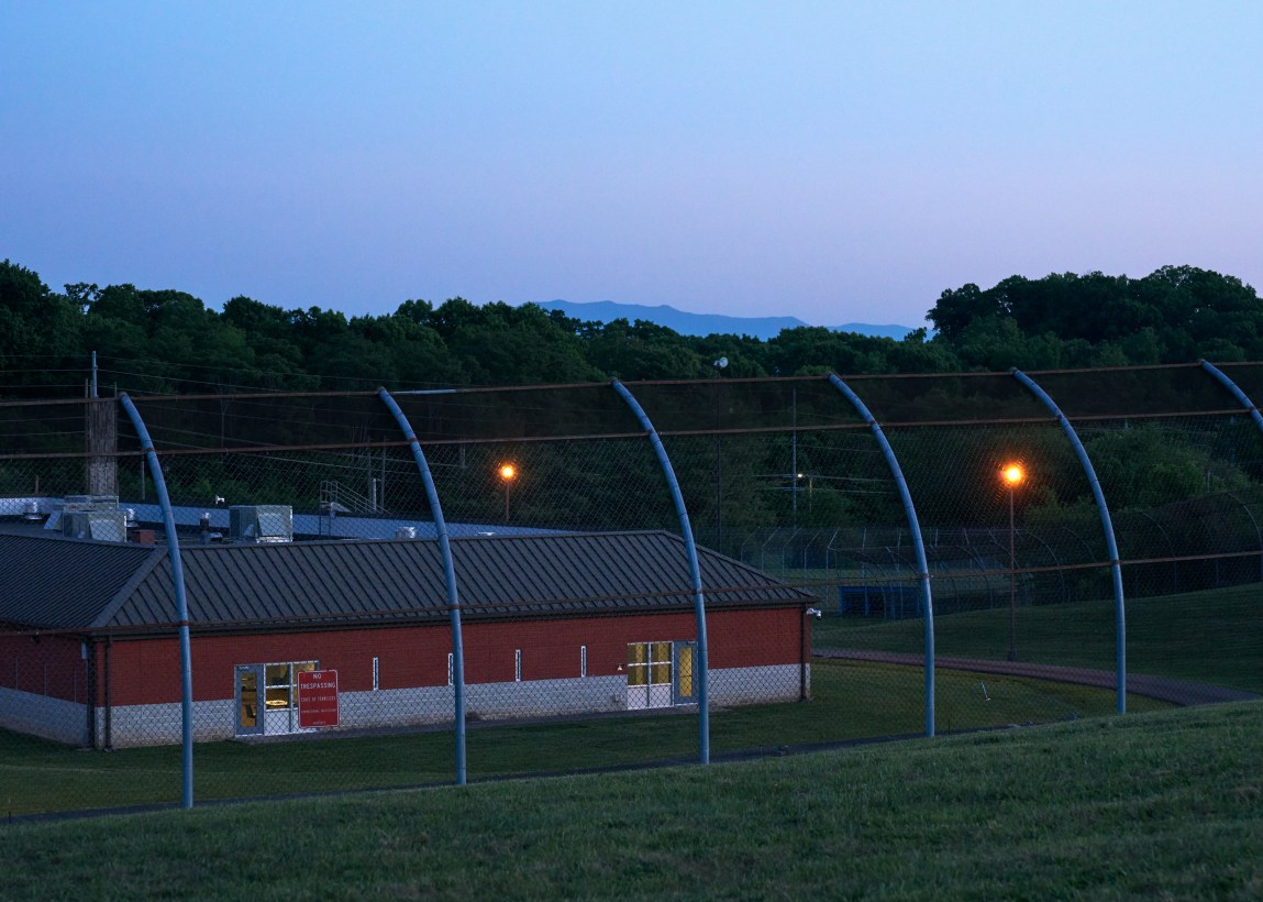 The exterior of a building surrounded by high fencing, with forests and mountains in the background, at dusk.