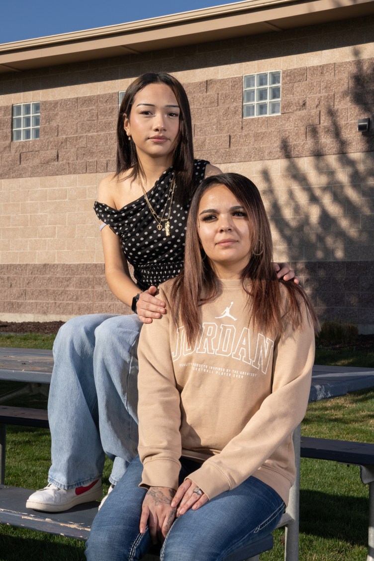 A teenage girl with long brown hair, wearing gold necklaces, a polka-dot black shirt and jeans rests her hands on her mother’s shoulders. Her mother also has long brown hair and is wearing a tan sweatshirt and jeans. They’re seated on a picnic table near a large tan building.