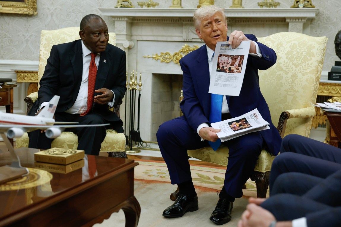 President Donald Trump sits in a gold patterned arm chair holding up a printed article to a crowd, next to South Africa President Cyril Ramaphosa, who is looking at him. Both men are in suits. 