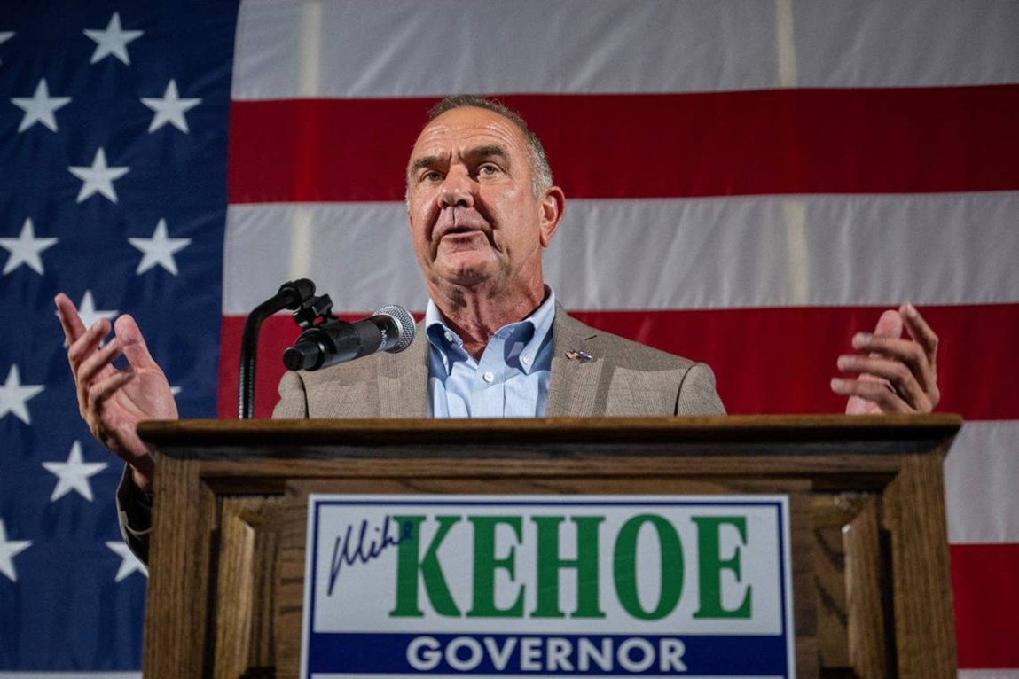 A man speaks at a podium during a campaign event, gesturing with his hands in front of a large American flag. A sign mounted on a wooden lectern reads “Mike Kehoe governor.”