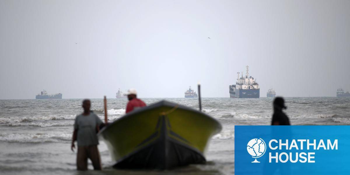 Three men bring a fishing boat into shore, while in the background large ships lie at anchor in the Strait of Hormuz.