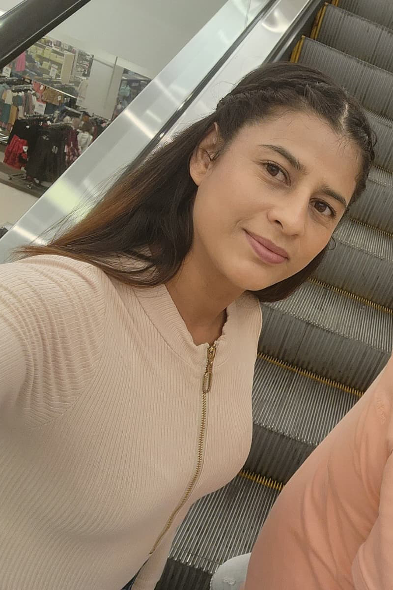 A woman with long dark hair and a tan shirt poses for a selfie on an escalator.