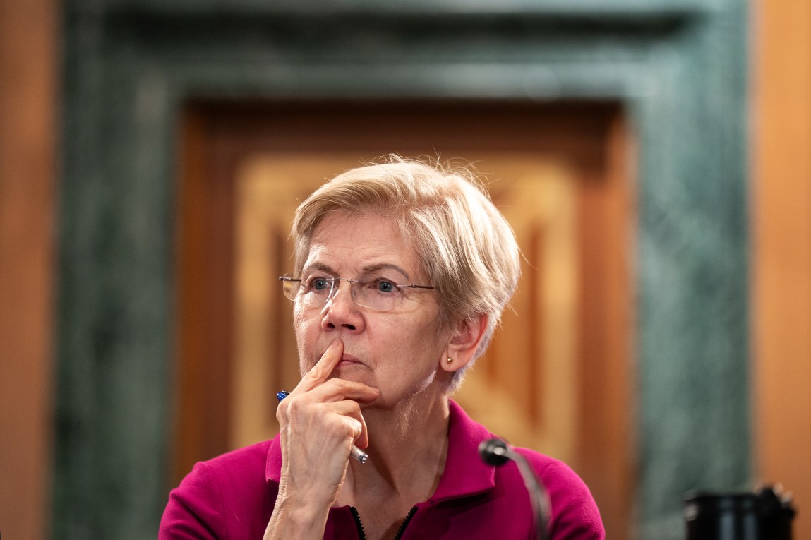 A woman with short blond hair and glasses looks off camera with her index finger over her mouth. She is framed by concentric rectangles in the background.