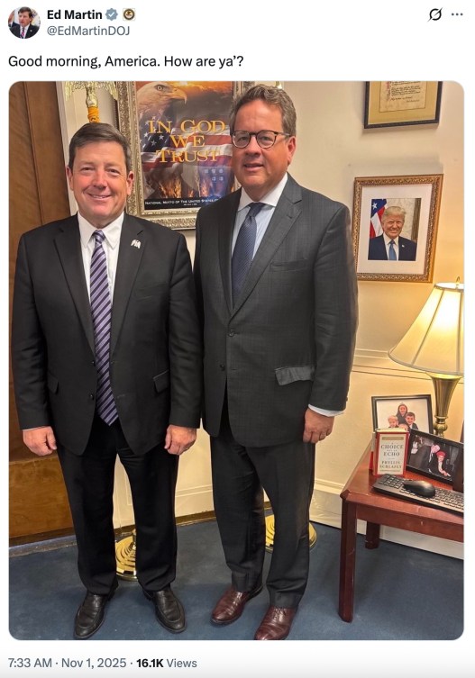 Two men wearing dark suits stand in a room with a blue carpet, white walls and a picture of President Donald Trump in the background.