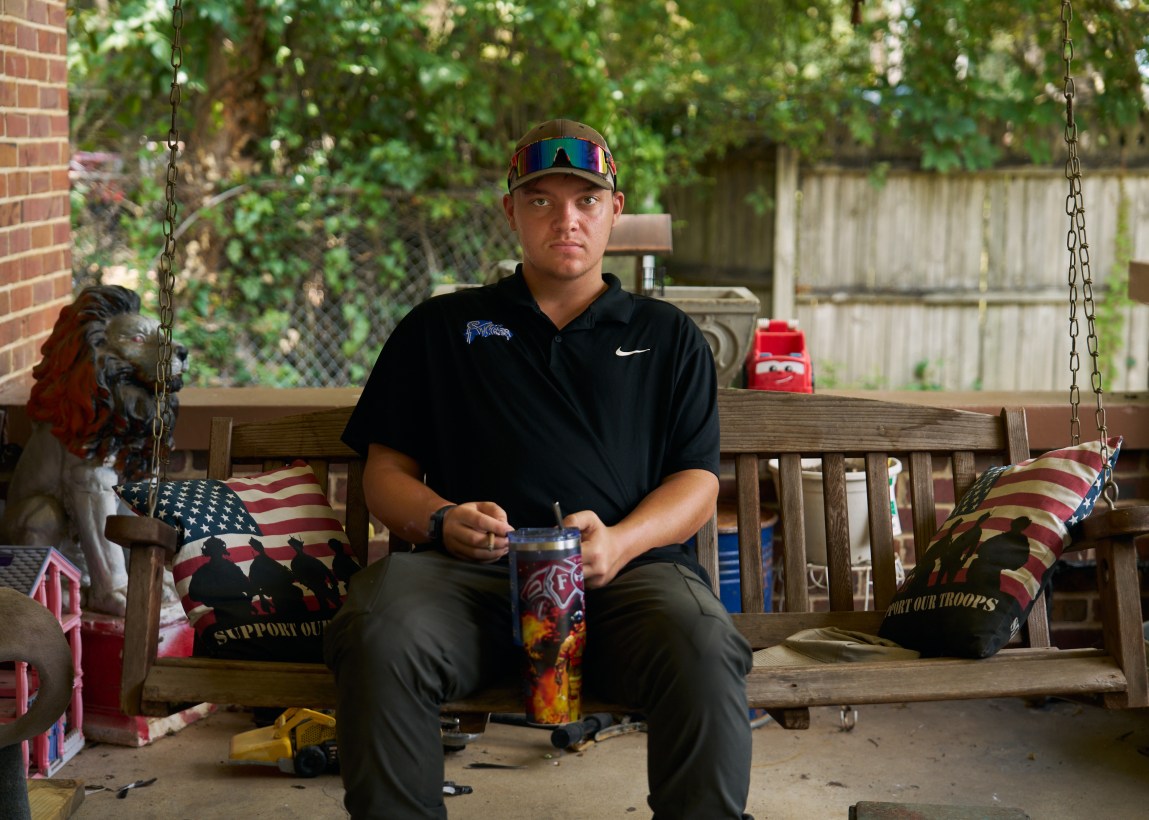 A young man sits on a porch swing looking at the camera with a serious expression. He is holding a cigarette in one hand and a water bottle in the other. On either side of him are pillows with American flags, silhouettes of soldiers and the words “Support Our Troops.”