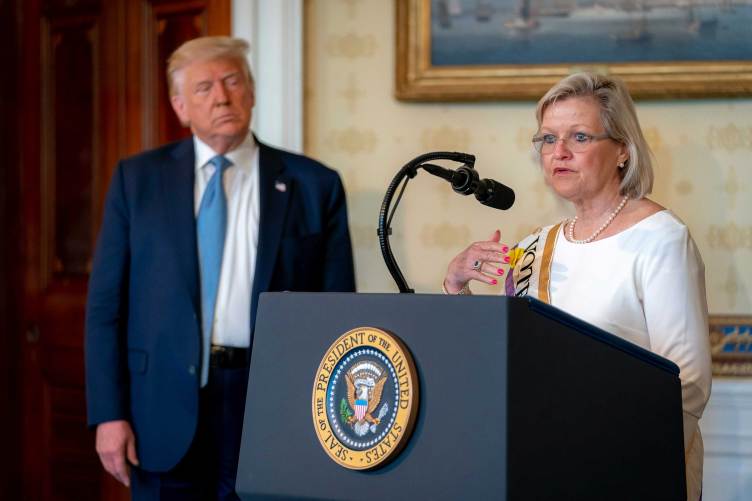 A woman wearing glasses, a white dress and a sash that reads, in part, “vote” speaks into a microphone at a podium bearing the seal of the president of the United States, while President Donald Trump watches from off to the side in a suit and blue tie.