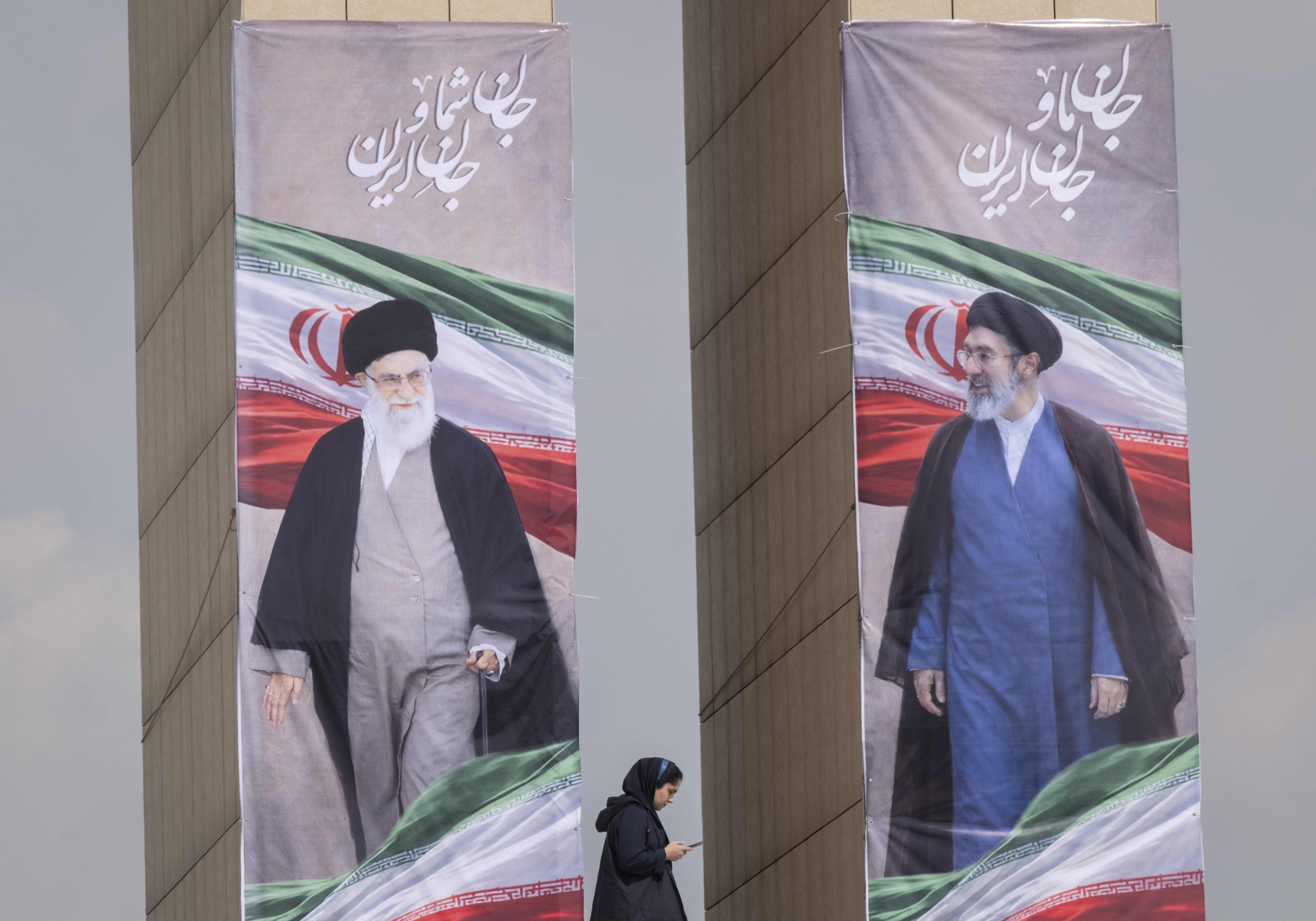 A young Iranian woman uses her cell phone while walking under portraits of the late Supreme Leader Ayatollah Ali Khamenei and the new Supreme Leader Ayatollah Mojtaba Khamenei during a flag ceremony marking Iran's Islamic Republic National Day in the Abbasabad Cultural and Tourist Area in central Tehran on April 1, 2026. This event takes place amid U.S.-Israeli military operations in Iran. Iranians voted in favor of the Islamic Republic regime in a referendum forty-seven years ago. (Photo by Morteza Nikoubazl/NurPhoto via Getty Images)