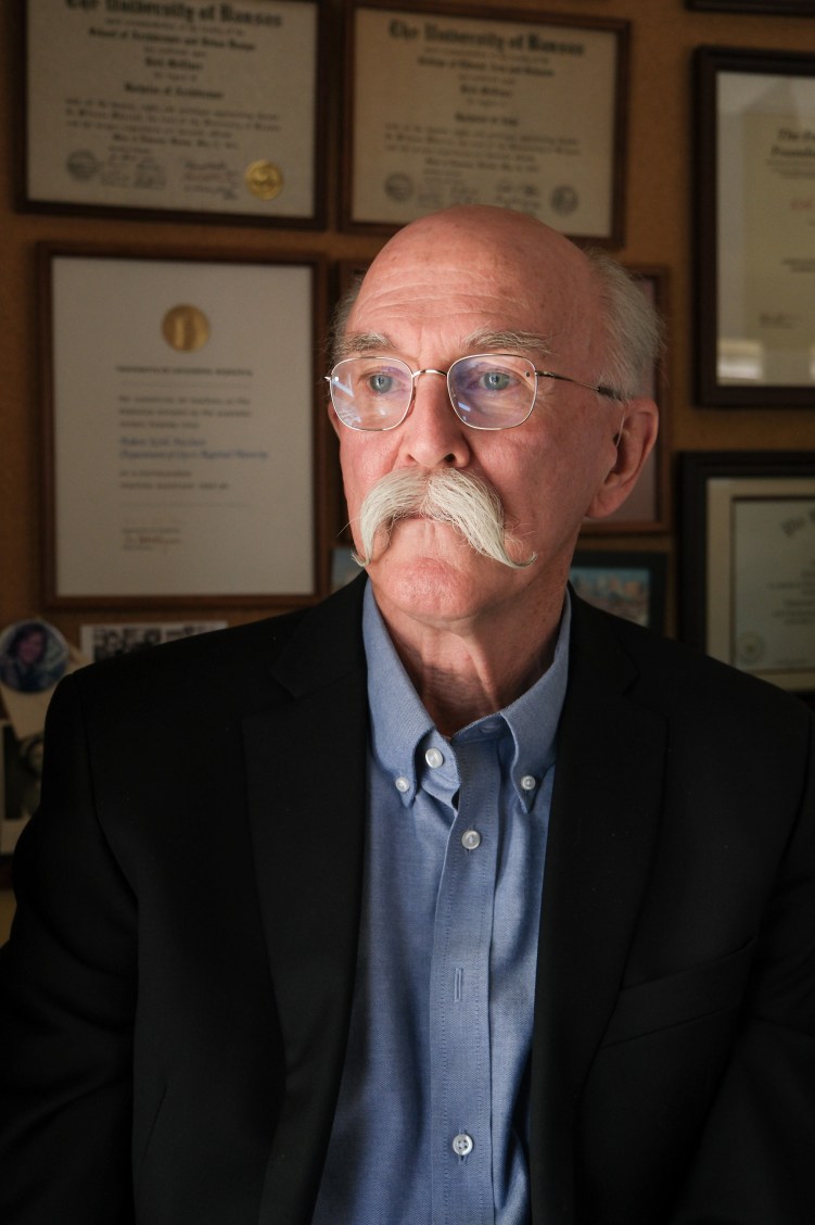 A man with blue eyes, white hair, silver-rimmed glasses and a large white mustache wears a black blazer and blue button-down shirt. He is in front of a grid of framed certificates and diplomas and looks off camera.