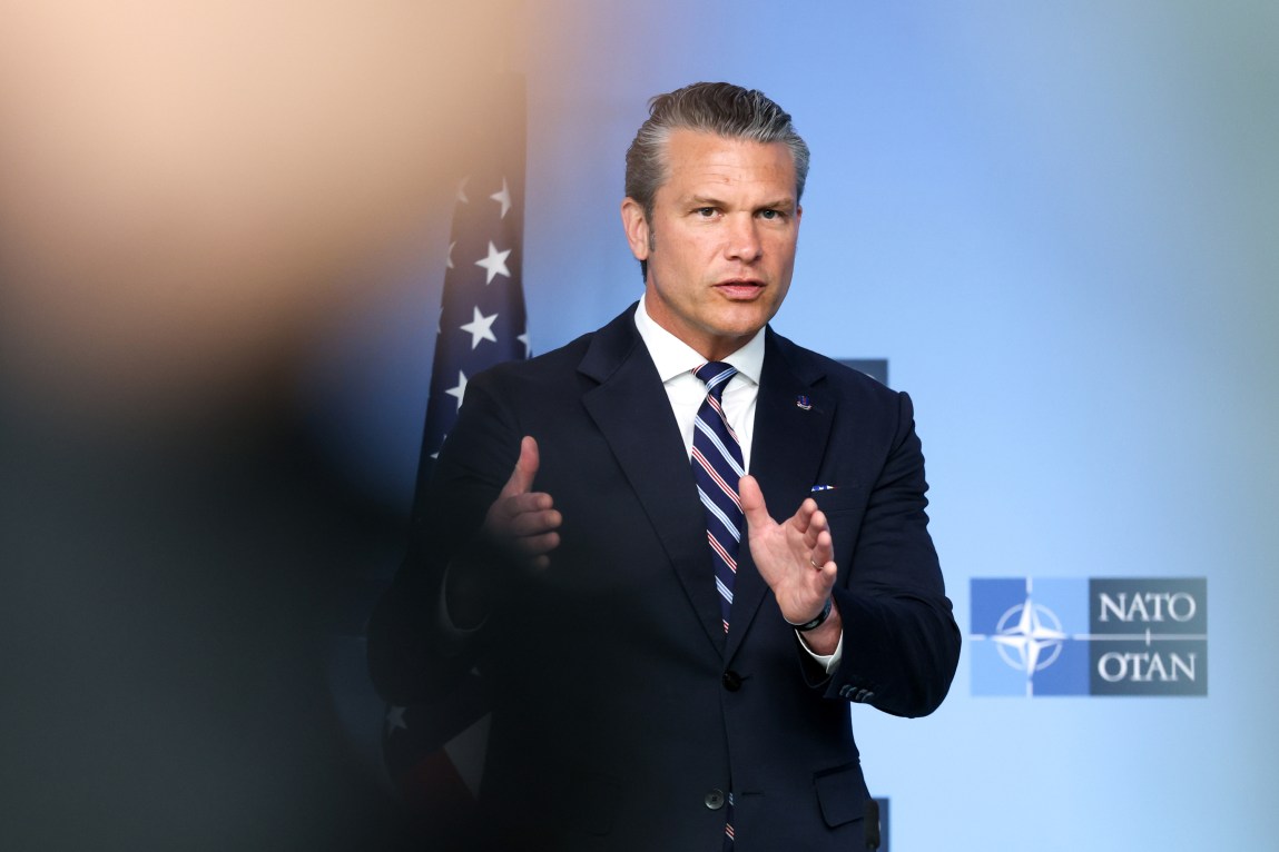 A man in a suit and tie gestures with his hands while speaking in front of an American flag.