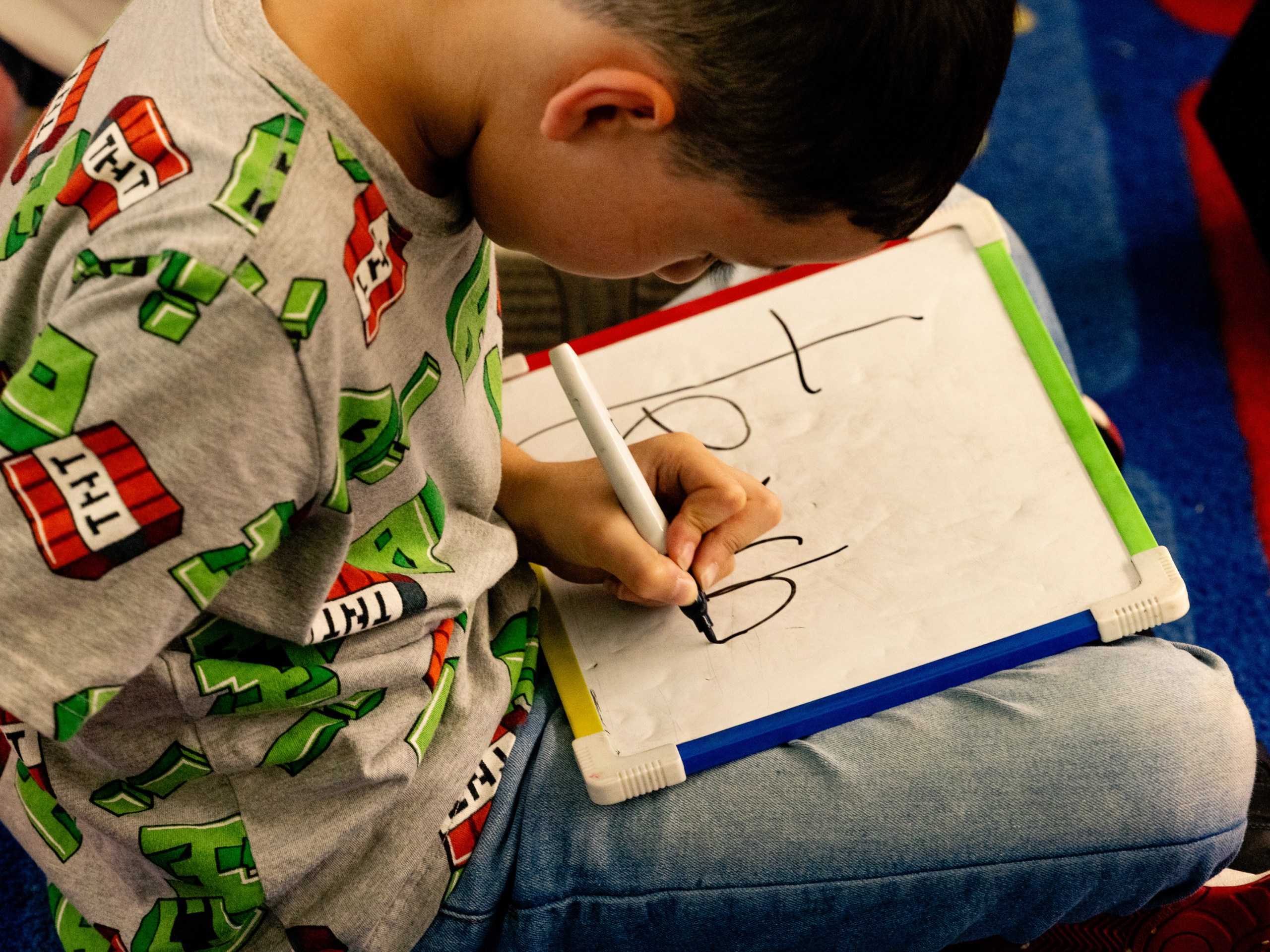 A young student writes on a small whiteboard while sitting cross-legged on the floor.