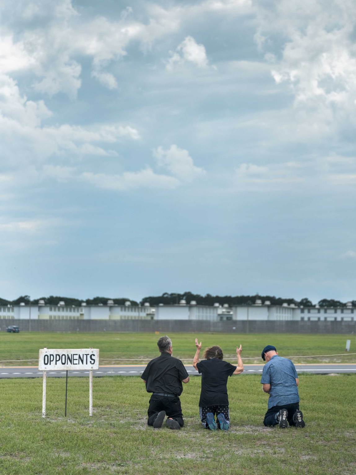 Three people are seen from behind, kneeling on a grassy lawn facing away from the camera toward a distant, low-profile facility with white buildings and a perimeter fence. To the left of the group, a small white sign on posts reads “Opponents” in black capital letters. The center individual has both hands raised in the air against a large expanse of cloudy sky.