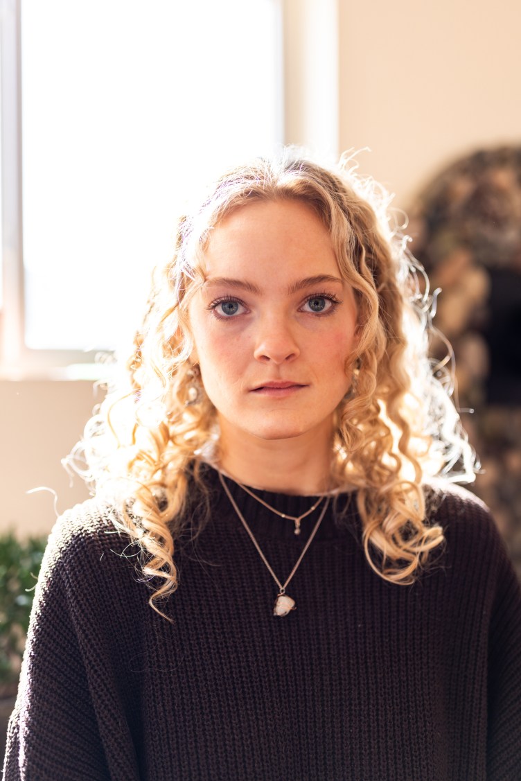A head-and-shoulders portrait of a woman in her 20s, looking at the camera with a neutral expression, lit dramatically.