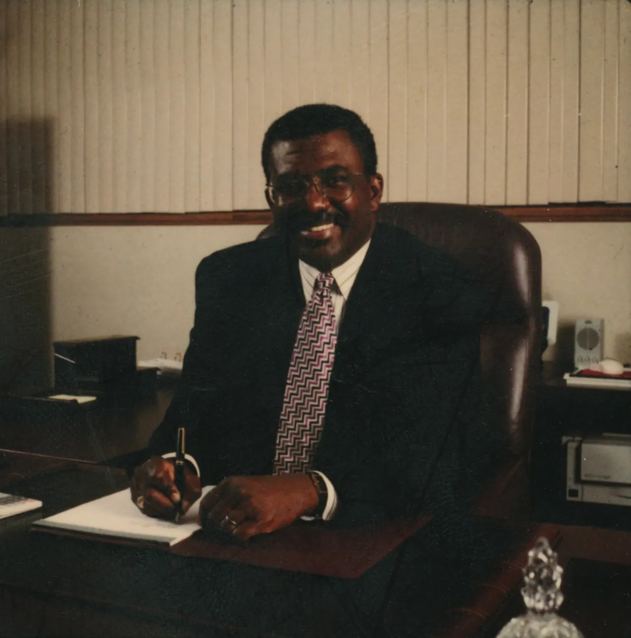 A man sits smiling at a desk wearing a suit. He is holding a pen and writing on a pad of paper.