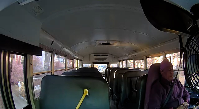 The interior of a school bus. At the front, a man sits in the driver’s seat with his eyes closed and his hands clasped in his lap.