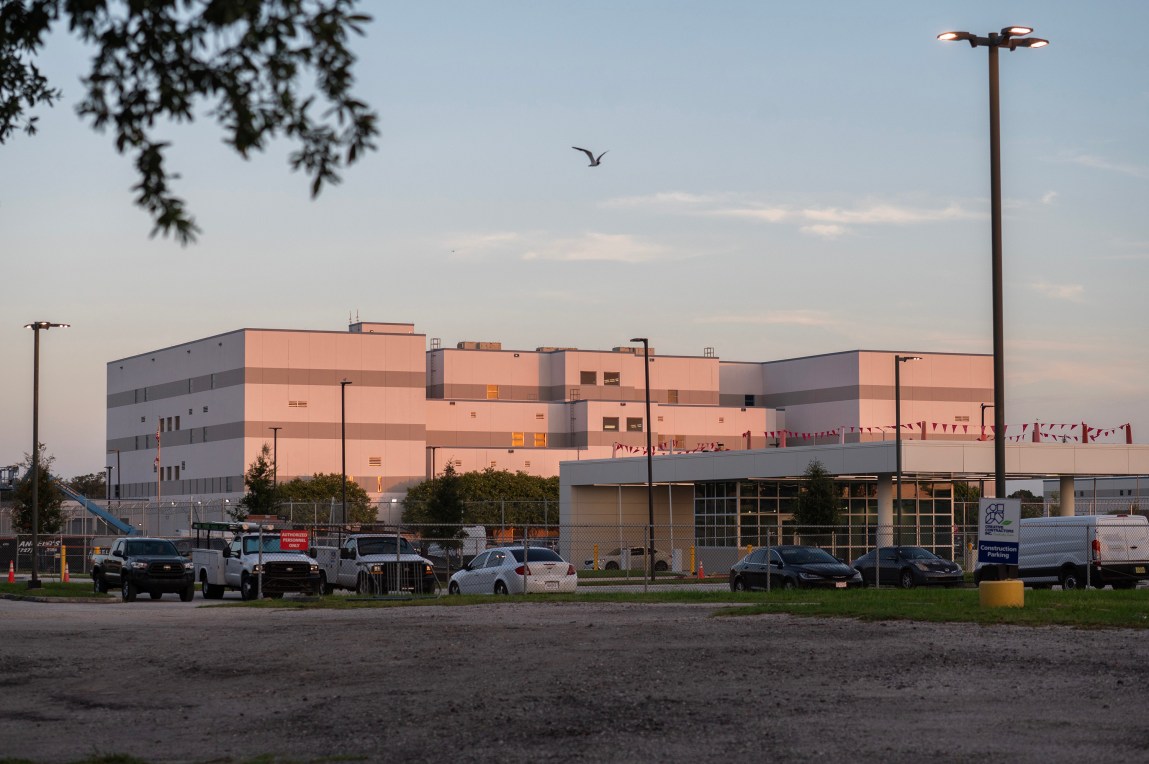 A large, modern multistory jail facility illuminated by warm evening sunlight behind a fenced parking lot.