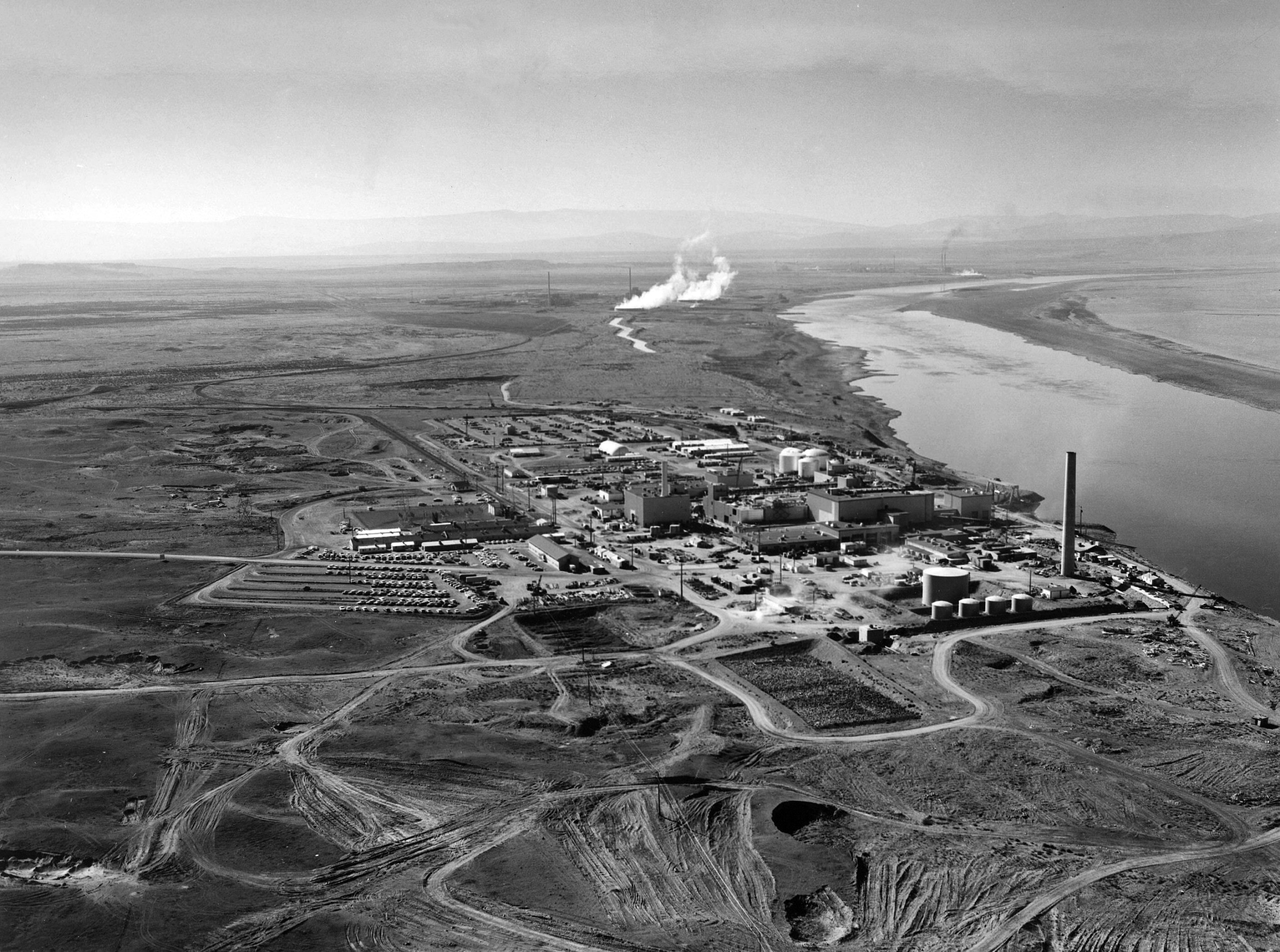 A black-and-white aerial photo of a plant with a tall smokestack and many big and small buildings. It sits in a dry environment next to a wide river.