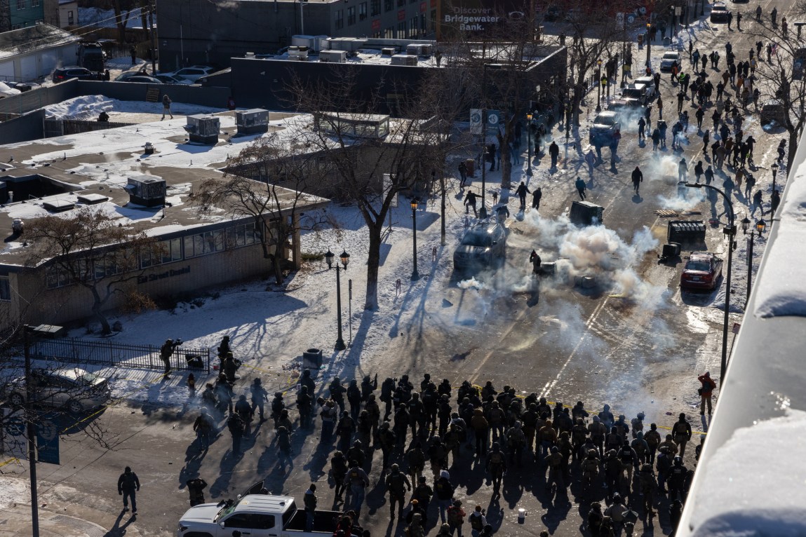 An overhead view of a mass of people in tactical vests standing behind a line of police tape and facing an urban street. Several clouds of gas are puffing up from the pavement as people in street clothes move away from the agents. Two dumpsters and multiple trash bins have been overturned in the roadway.