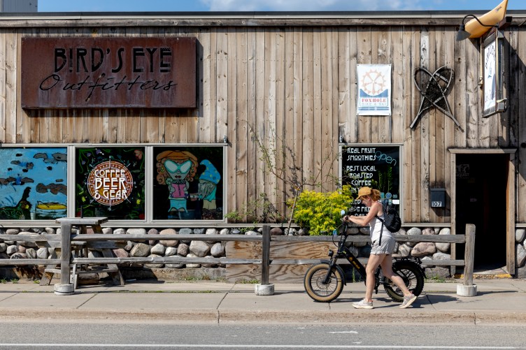 A woman wearing a tan cap, white tank top, shorts and a backpack pushes a black electric bike along a sidewalk in front of a rustic wooden storefront. The building features a metal sign reading “Bird’s Eye Outfitters” above windows decorated with artwork and text advertising coffee, beer, gear and smoothies.