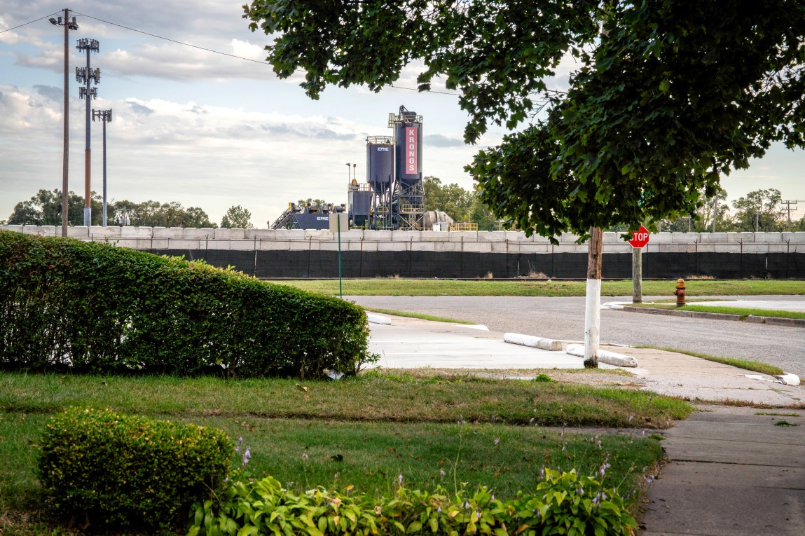 A large industrial structure with the word Kronos written on it overlooks a neighborhood of green lawns.