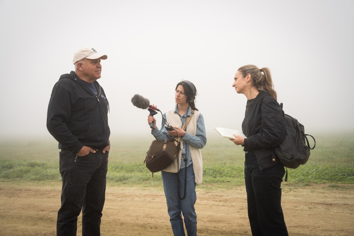 A man being interviewed in a foggy field by two women, one holding a microphone and the other a notebook.