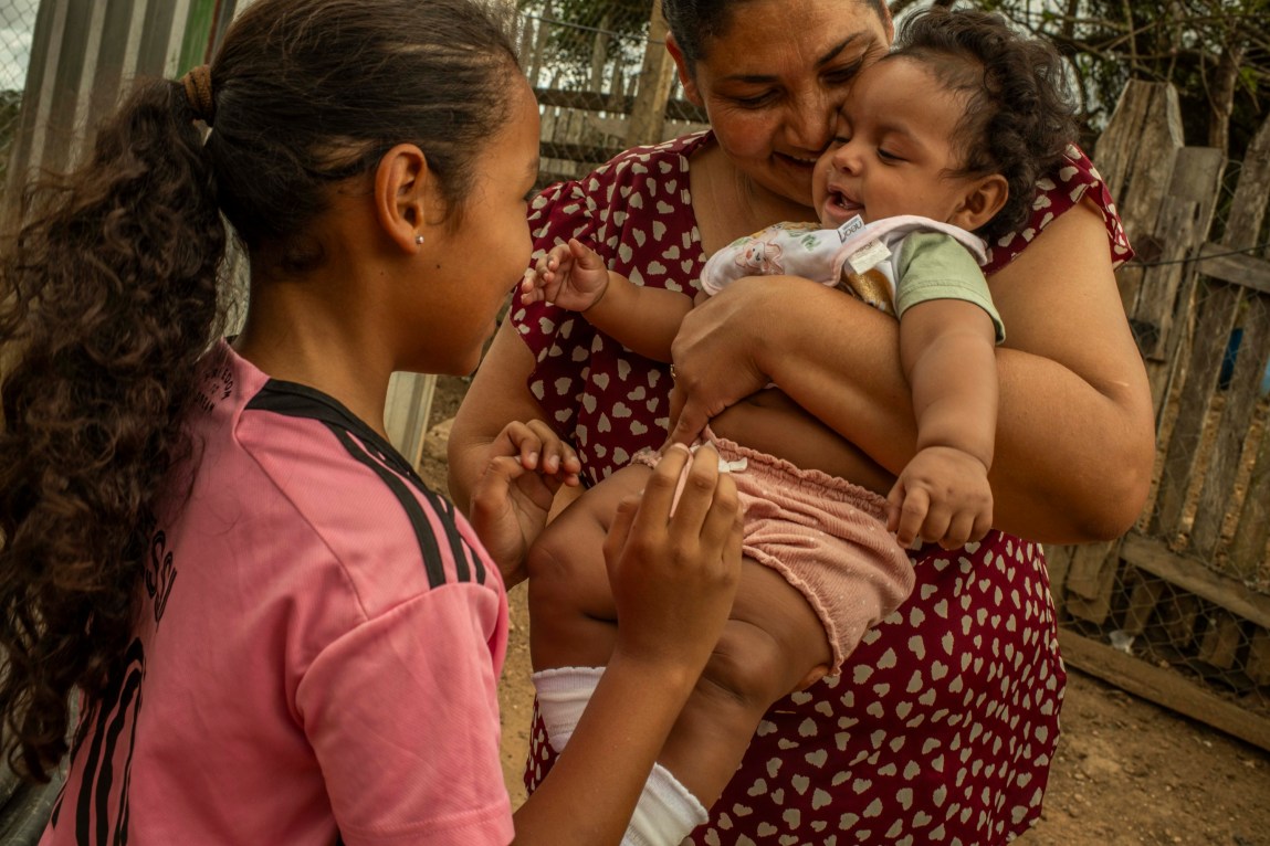 A woman wearing a red blouse with pink hearts cuddles an infant in her arms as a young girl wearing a pink soccer jersey makes the baby laugh.