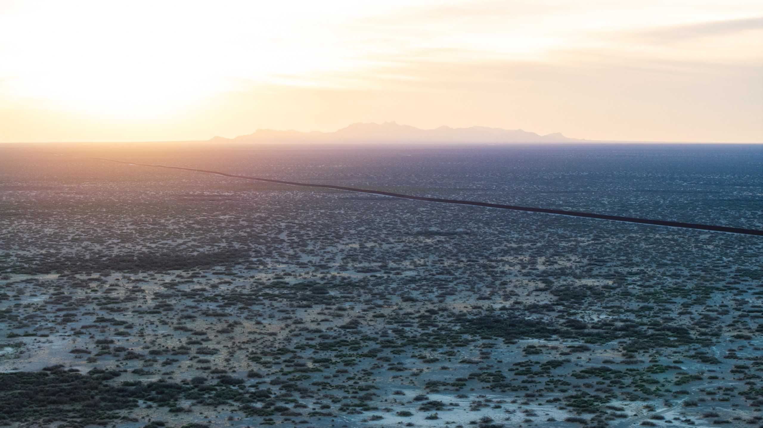 A vast desert landscape, showing a thin U.S. border wall with Mexico stretching off into the setting sun.