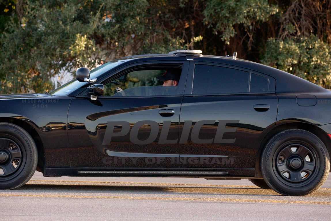 An officer’s baseball cap and the top of his face are visible above the partly lowered driver’s-side window of a black Dodge Charger that says “Police” and “City of Ingram” across its doors.