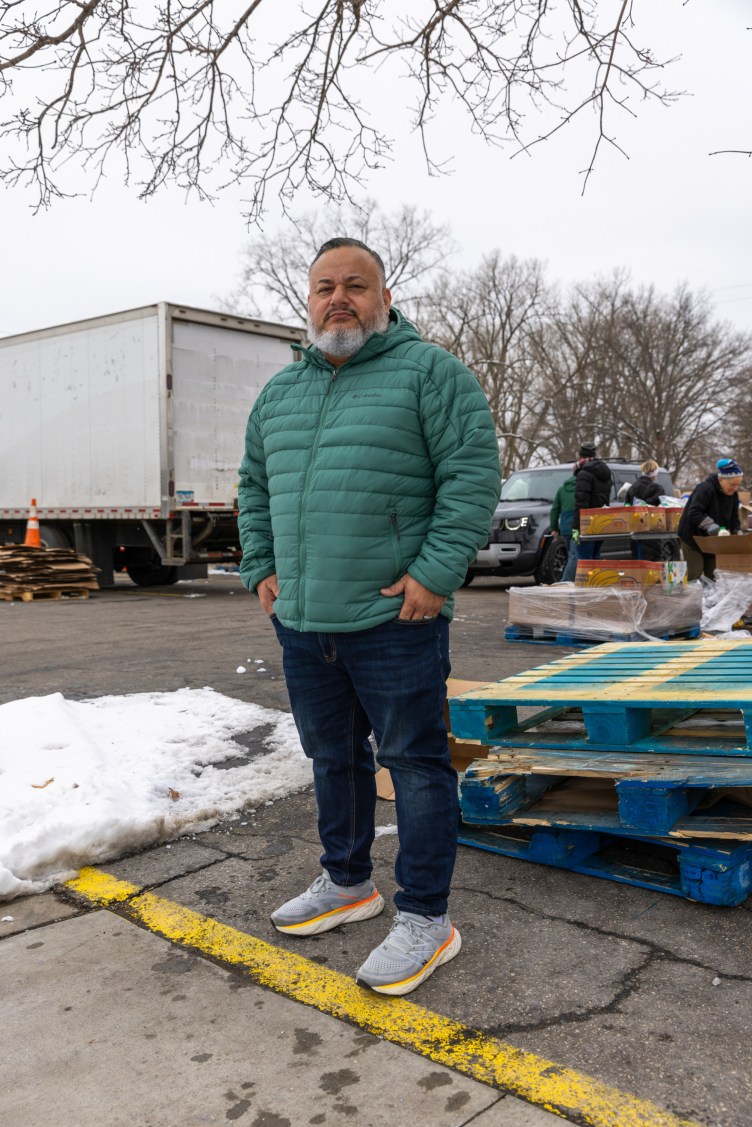 A man stands with his hands in his pockets looking at the camera, in a snow-covered parking lot. In the background, people load boxes of food into cars.