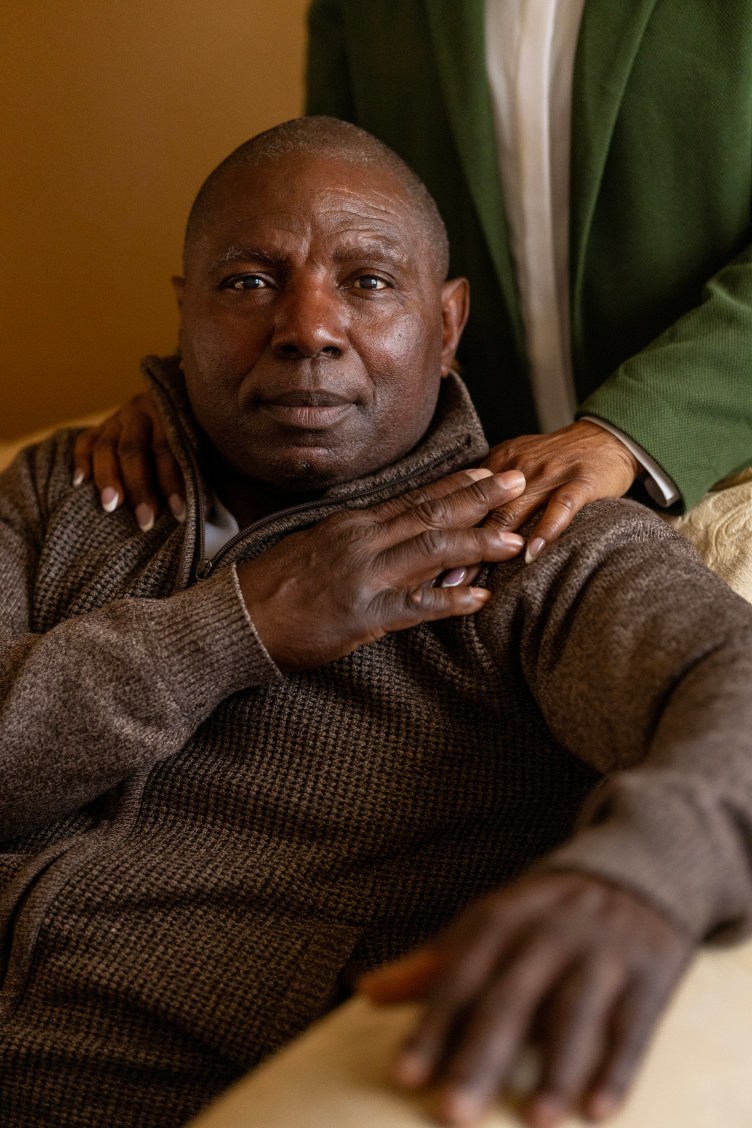 A man in a brown sweater sits on a chair looking at the camera. A person behind him rests their hands on his shoulders, and he has his hand placed on top of theirs.