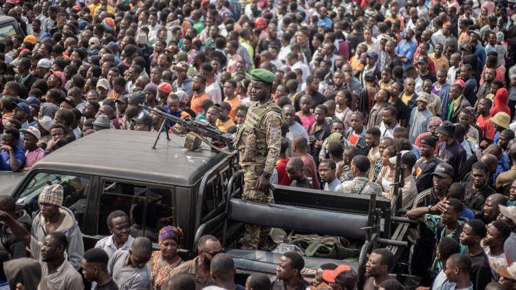 A soldier stands on a pick up truck holding a maching gun at a large gathering.