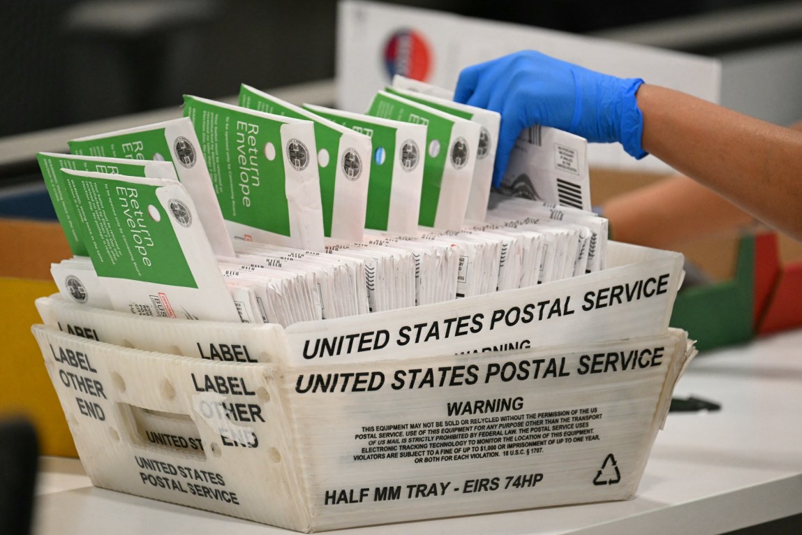 A hand with a blue rubber glove reaches toward ballots labeled “Return Envelope” in a box labeled “United States Postal Service.”