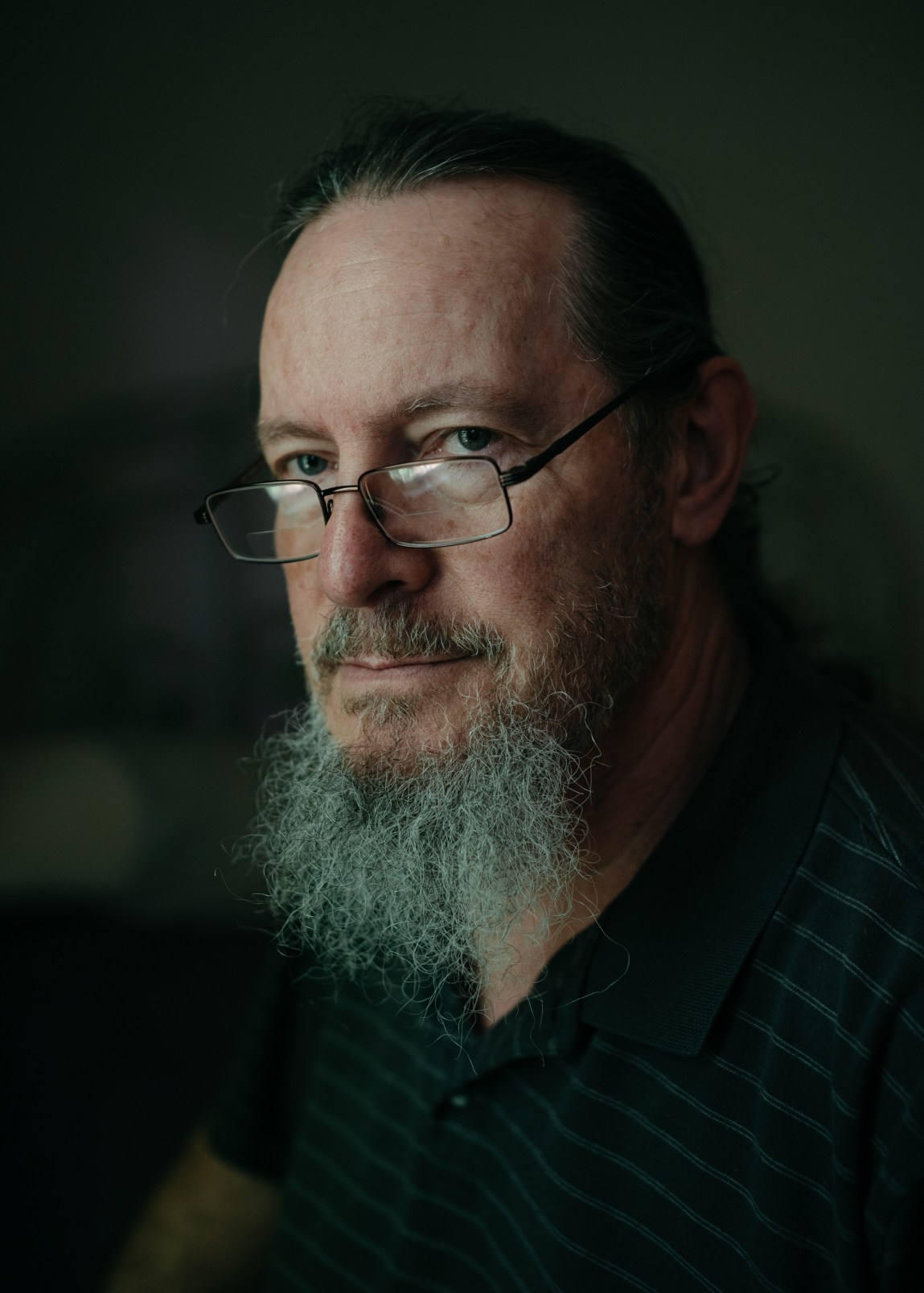 A man with glasses and a gray beard wears a striped polo shirt. He sits in a dark room.