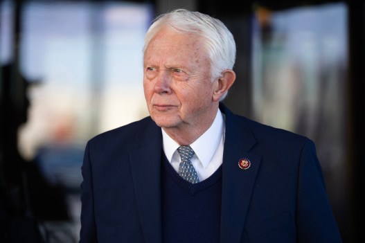 A man wears a navy blue suit jacket, a matching dark sweater and a gold-and-blue patterned tie over a white collared shirt. A pin is attached to his left suit lapel.