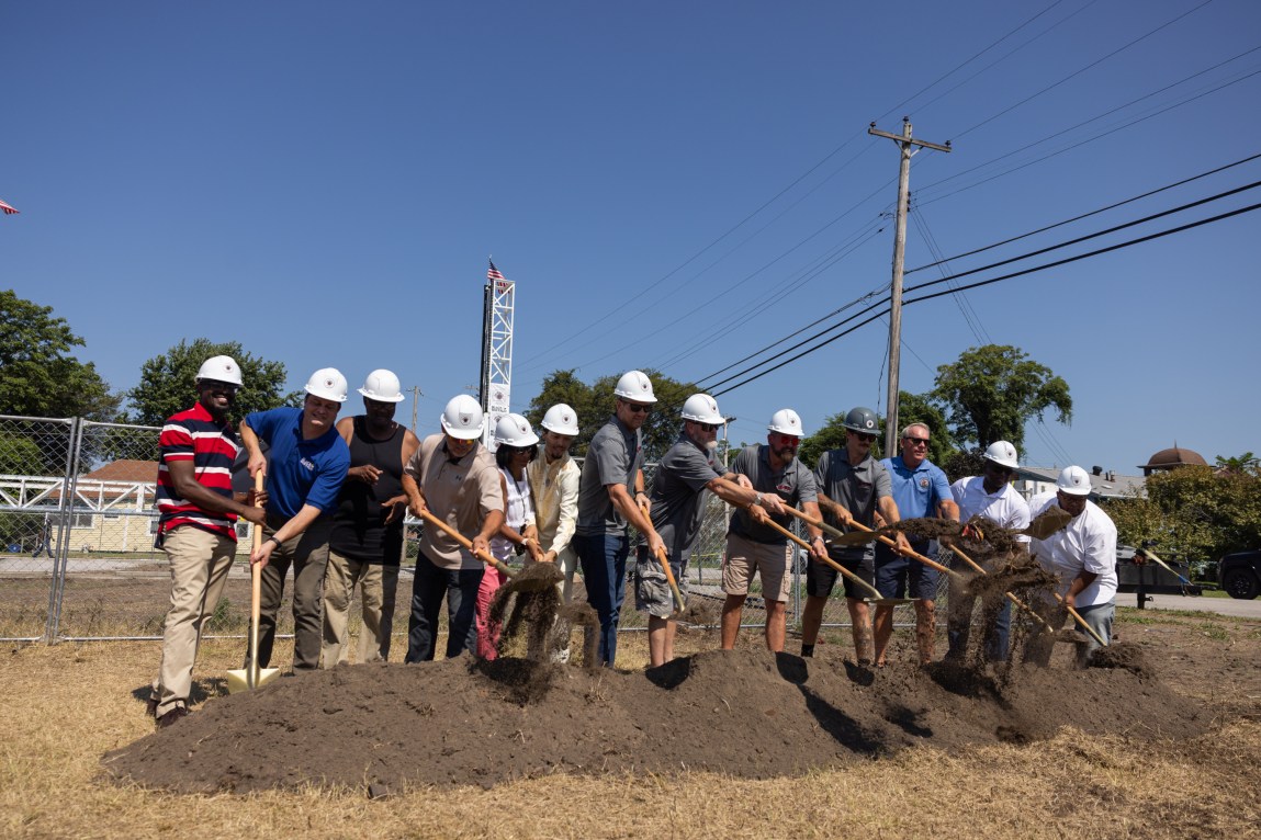 Thirteen people with hard hats stand in a row and shovel a pile of dirt outdoors. Power lines and a structure that looks like a tower, part of a huge 3D printer, are in the background.