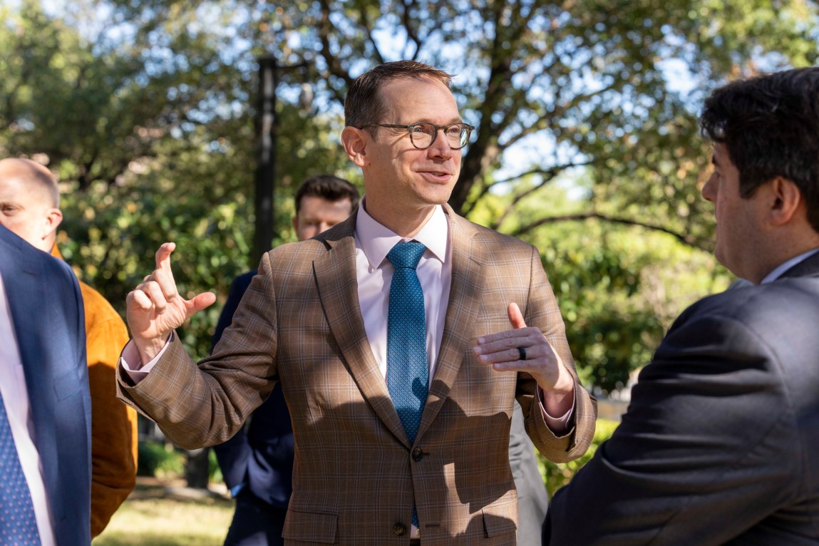 A man wearing glasses, a brown suit and blue tie smiles as he gestures with his hands. He is standing outside in front of trees with other people wearing suits.