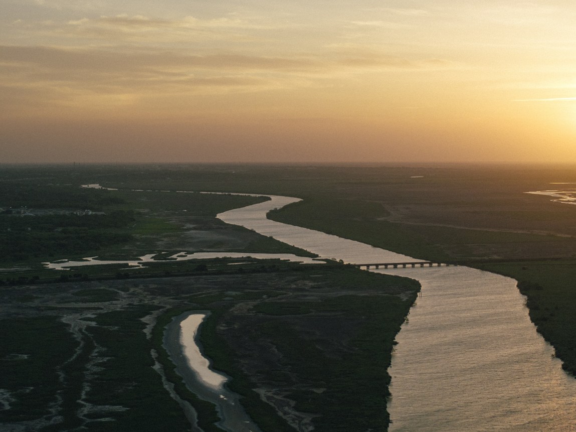 An aerial landscape photo shows a winding river snaking through dark marshlands and green terrain toward the horizon under a glowing sky at sunset. A low bridge crosses the river in the midground, and calm waters reflect the warm ambient light.