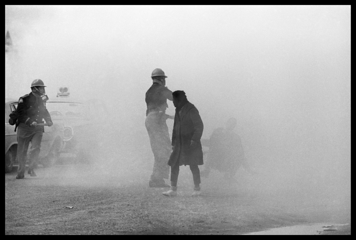A black-and-white photograph of a scene that is obscured by a cloud of tear gas. Two police officers and one other man are visible. Another person is barely visible in the haze as they fall to the ground.
