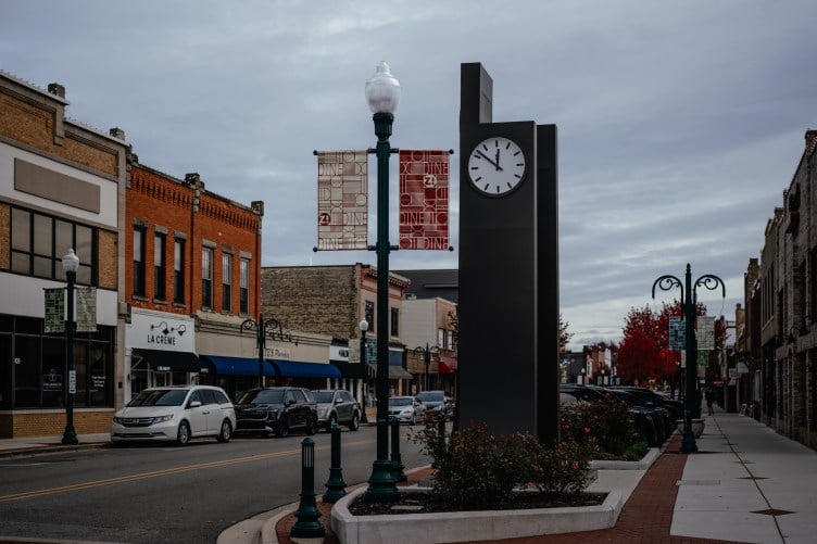 A small downtown area with cars parked along both sides of a two-lane street, storefronts and a large clock along the sidewalk.