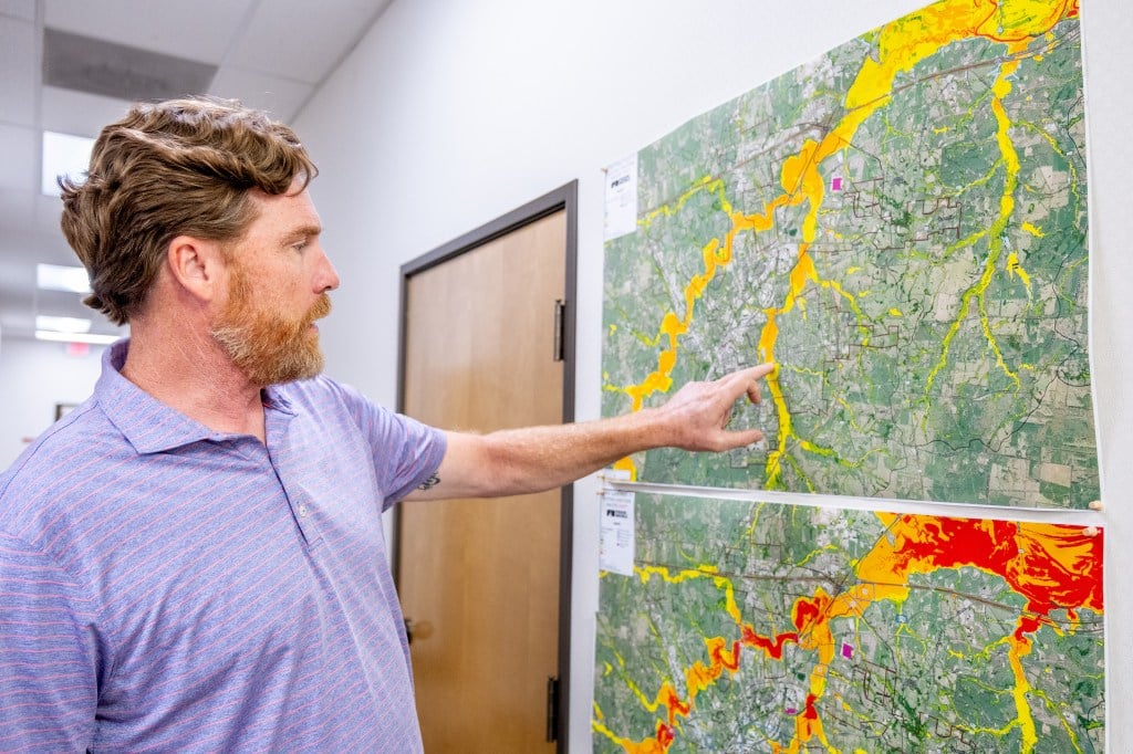 A man with brown hair and a short beard wearing a purple polo shirt points to a map hanging on an office wall.