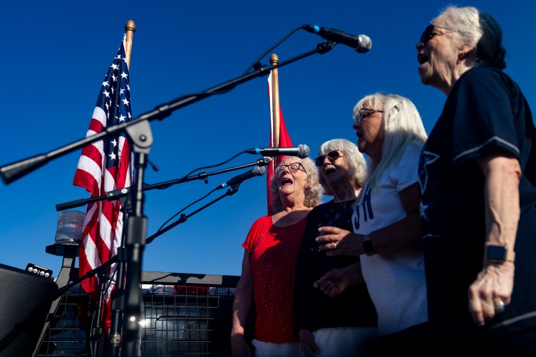 Four women stand side by side singing into microphones outdoors during a daytime event. Flags flank them in the background against a clear, deep blue sky.