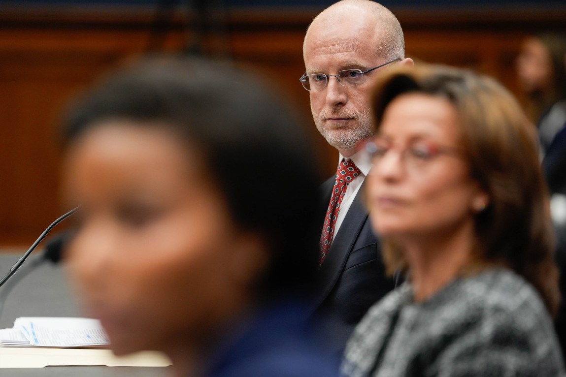 A man in a suit and glasses is in focus in the background. In the foreground are two women out of focus.