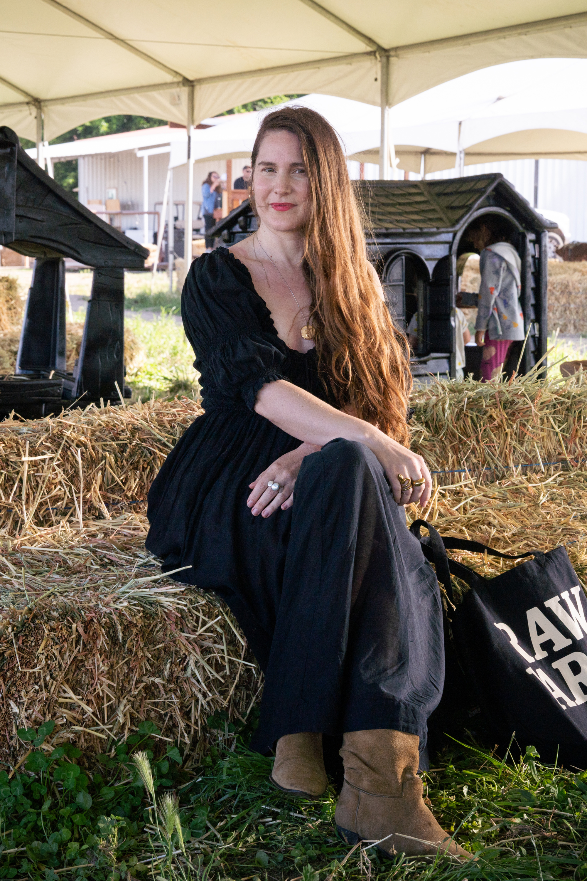 A woman in a black dress sits on hay bales under a large white tent, with a black Raw Farm tote bag resting beside her. Other people and children’s play structures are visible in the grassy background.